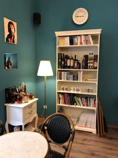 Weißer Regalständer mit Büchern, Flaschen und Dekoration, daneben Tisch mit Lampe und Stuhl.