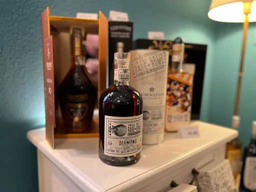 Dunkle Flasche mit Etikett auf weißem Regalbrett neben anderen Spirituosenflaschen und Kartons.