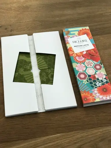 Weiße Verpackung mit grünem Teebeutel und bunte Packung mit Blumenmuster, Teebeutel sichtbar.